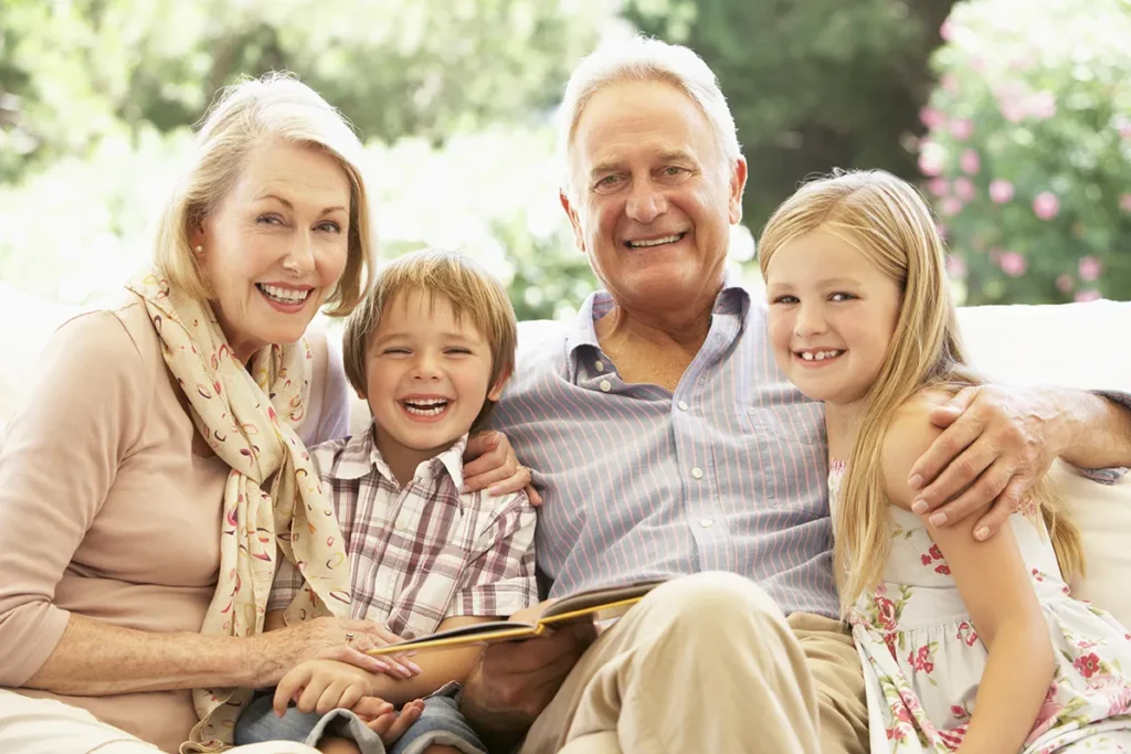 Portrait Of Grandparents Reading To Grandchildren On Sofa Smiling to Camera